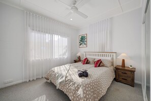 Een slaapkamer met een bed, twee nachtkastjes met lampen, een plafondventilator en een raam met dunne gordijnen.