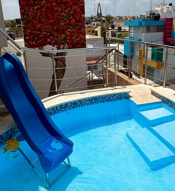 A swimming pool with a blue slide, surrounded by a metal fence, and a view of buildings in the background.