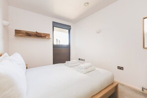 Une chambre à coucher avec un lit, des oreillers, une fenêtre avec des stores, une lampe fixée au mur et une petite étagère en bois sur laquelle est posé un objet décoratif.