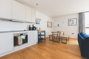 A modern kitchen with white cabinets, a wooden dining table, and a blue sofa. - Stylish Camberwell Two-Bed Retreat (London)