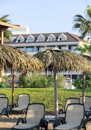 Outdoor seating with thatched umbrellas, a pool area, and a building with a tiled roof in the background. - Tui Blue Sherwood Belek (Serik)
