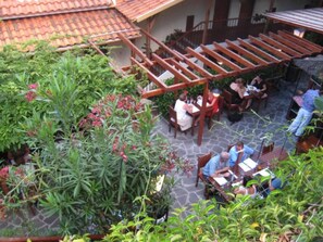 Un espacio al aire libre con un patio que cuenta con pérgolas de madera, mesas y sillas, rodeado de abundante vegetación y plantas en macetas.