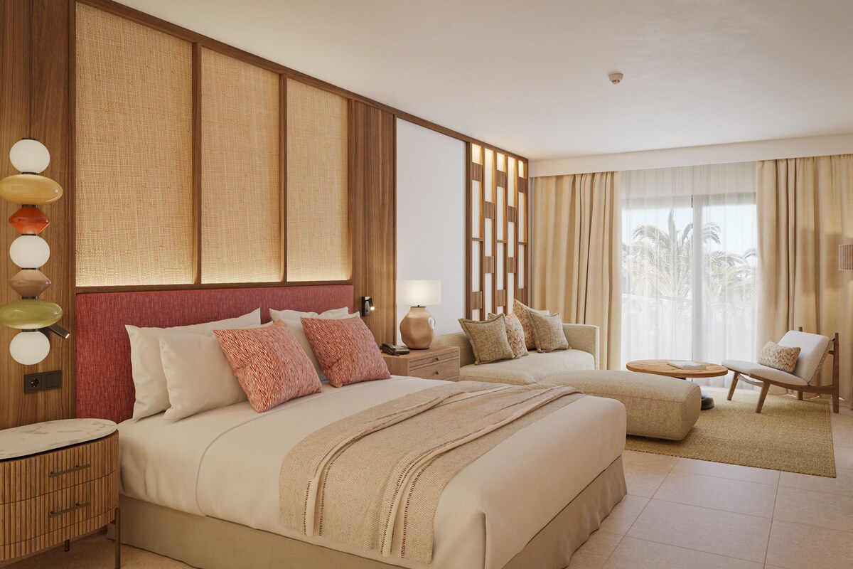 Una habitación de hotel con una cama grande, una mesita de noche, una lámpara, una silla y vistas a palmeras a través de la ventana.