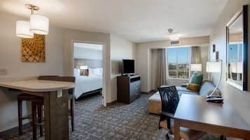A hotel room with a desk, chair, TV, and a sofa with a view of the city.
