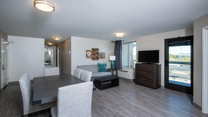 Quarto de hotel moderno com área de refeições, sofá, televisão e uma janela grande com vista.
