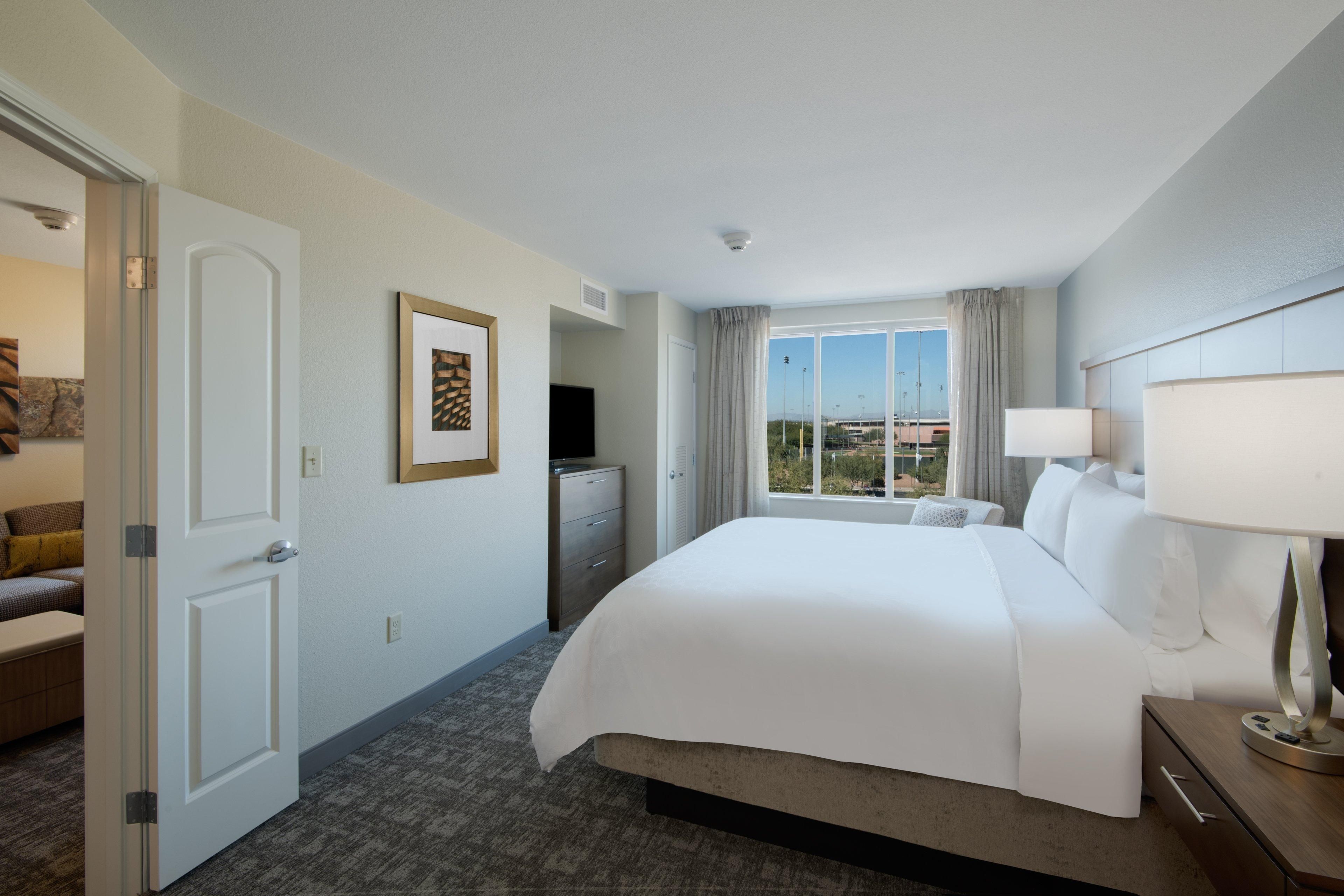 A hotel room with a large bed, a desk with a lamp, a TV, and a window with curtains.