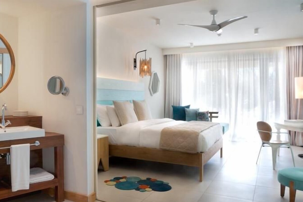 Habitación de hotel moderna con tocador de madera, cama con cabecera, ventilador de techo y vistas al exterior a través de amplios ventanales.
