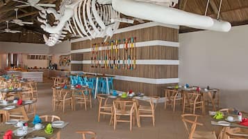 A restaurant with wooden chairs and tables, a large hanging skeleton decoration, and a wall with colorful bottles.