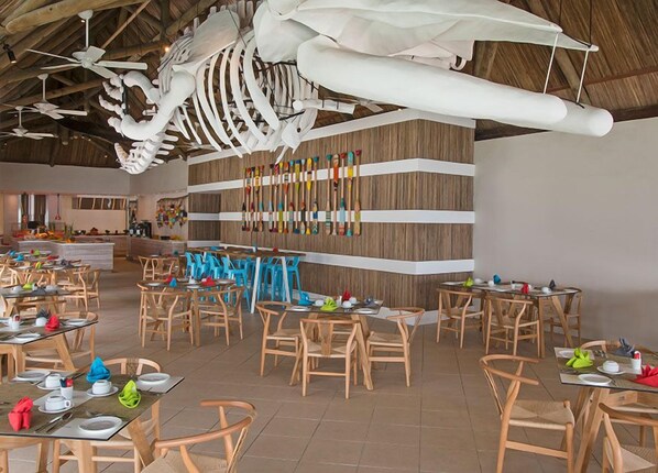 Un restaurante con sillas y mesas de madera, una gran decoración de esqueleto colgante y una pared con botellas de colores.