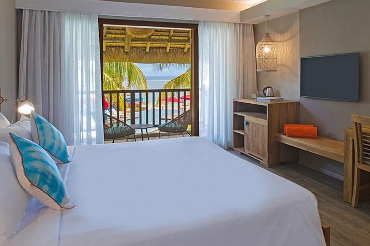 Habitación de hotel con cama, escritorio, silla, televisión y vista a una piscina y palmeras.