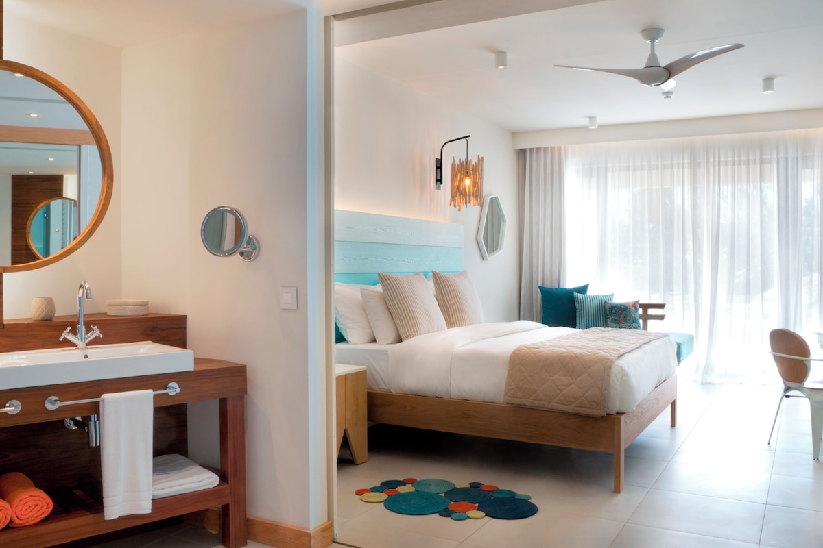 Habitación de hotel moderna con un tocador de madera, una cama grande con cabecera, un ventilador de techo y vistas al exterior a través de amplias ventanas.