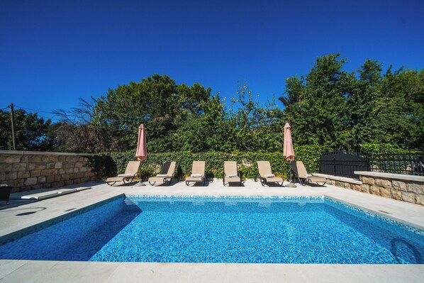 A rectangular swimming pool with clear blue water, surrounded by lounge chairs and umbrellas, set against a backdrop of trees and a stone wall. - Villa Franica (Dubrovnik)