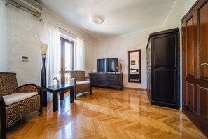 Una habitación con piso de parqué de madera, un armario de madera oscura, un televisor, un sofá de mimbre y una mesa con una lámpara.