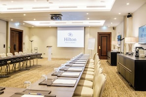 Un salón de conferencias con una larga mesa, sillas, una pantalla de proyector y una pantalla mostrando el logotipo de Hilton.