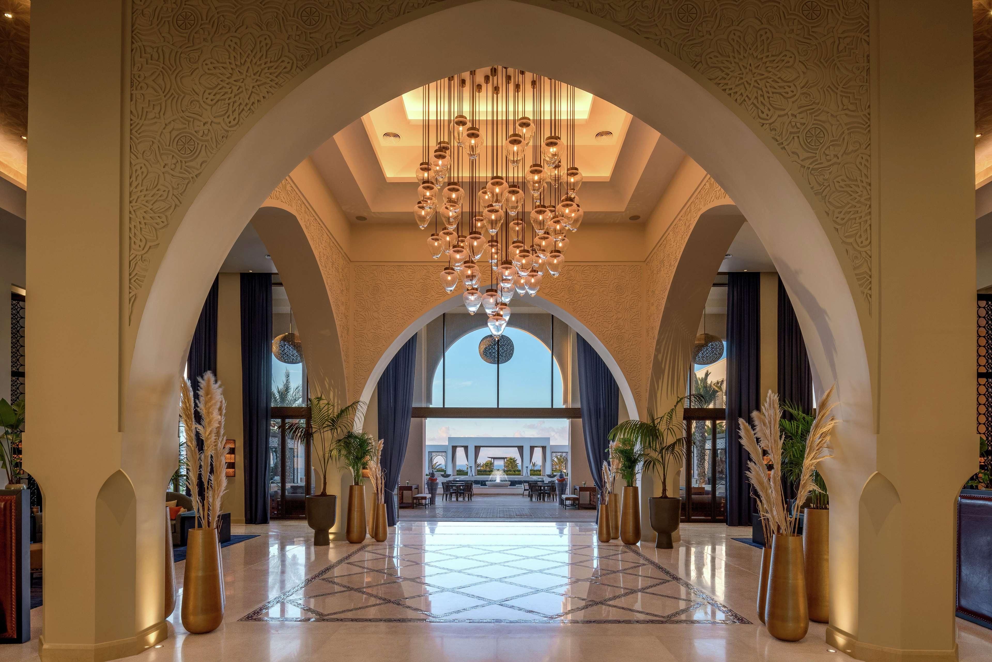 A spacious lobby with a high ceiling, ornate wall carvings, and a central chandelier.