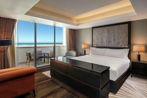 Une chambre d’hôtel moderne dotée d’un grand lit, d’un mur en verre donnant sur la plage, d’un coin repas et d’un espace salon séparé.