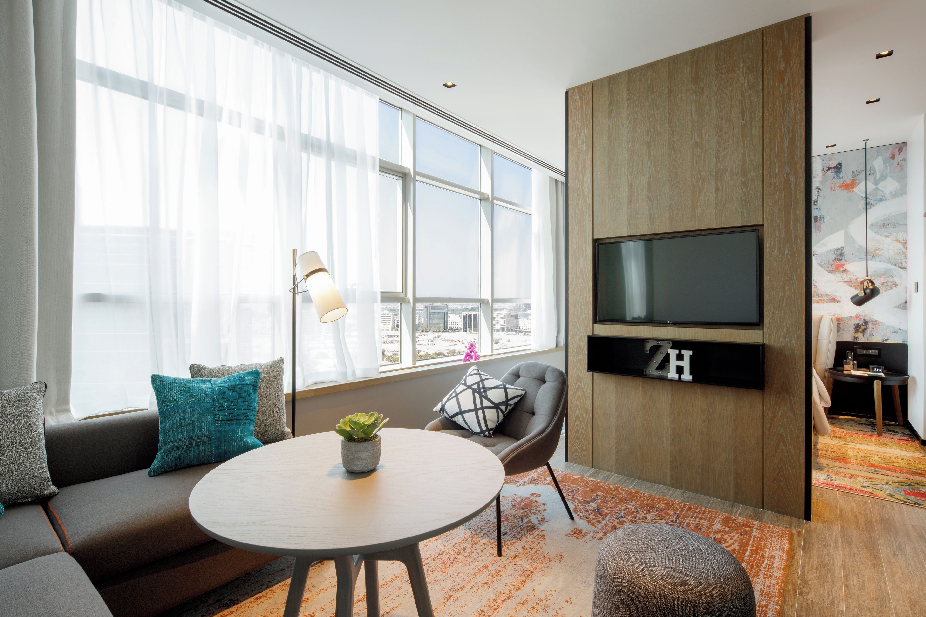 A modern living room with a sofa, a round table, a TV mounted on a wooden wall, and a large window with sheer curtains.