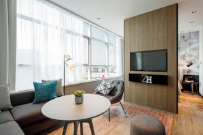 Una sala de estar moderna con un sofá, una mesa redonda, un televisor montado en una pared de madera y un amplio ventanal con cortinas translúcidas.