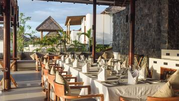 An outdoor dining area with wooden chairs and tables, set against a backdrop of a stone wall and a thatched-roof structure.
