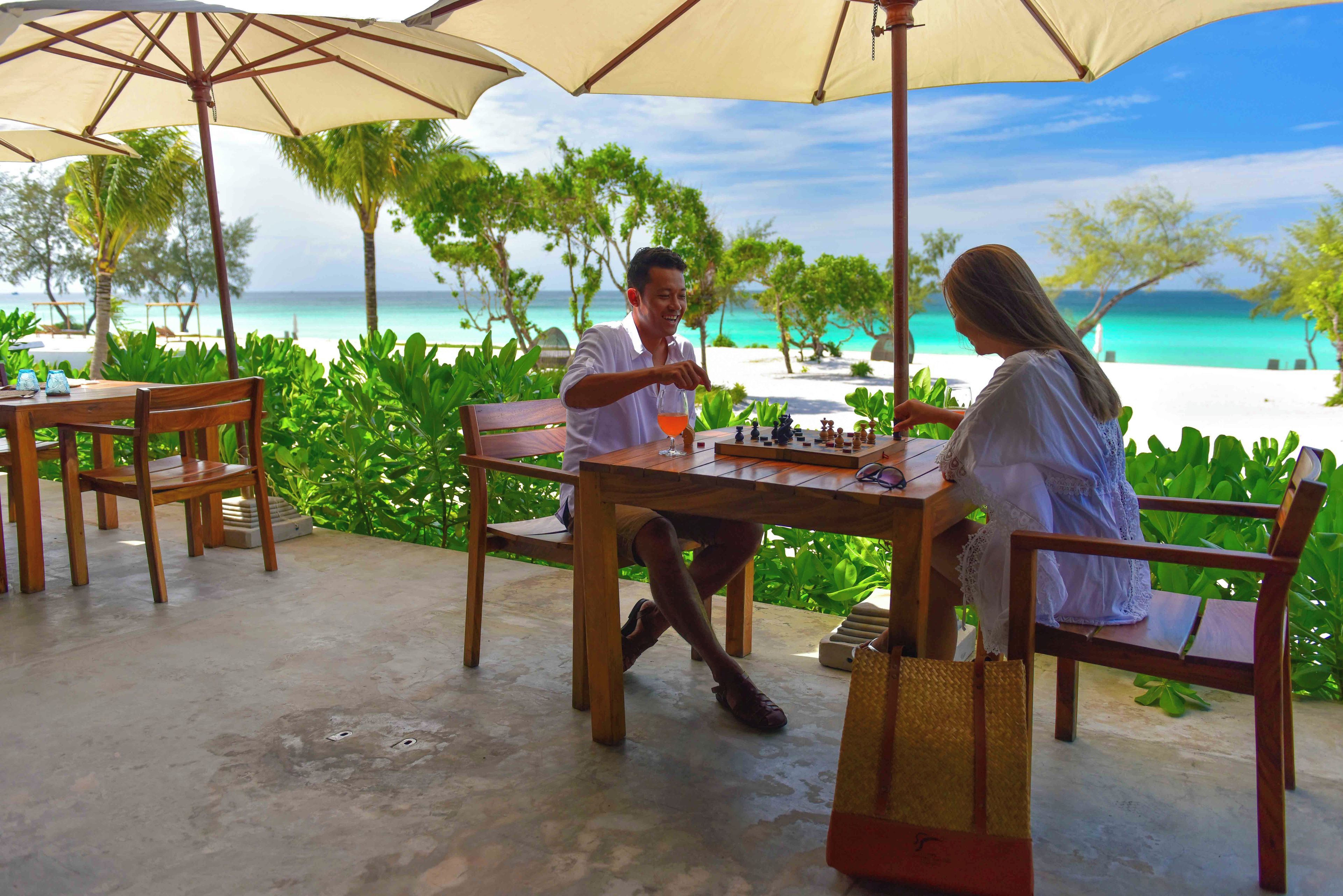 A wooden table with chess set, two chairs, and a table umbrella at a beachside location.