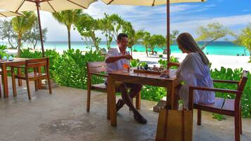 A wooden table with chess set, two chairs, and a table umbrella at a beachside location.