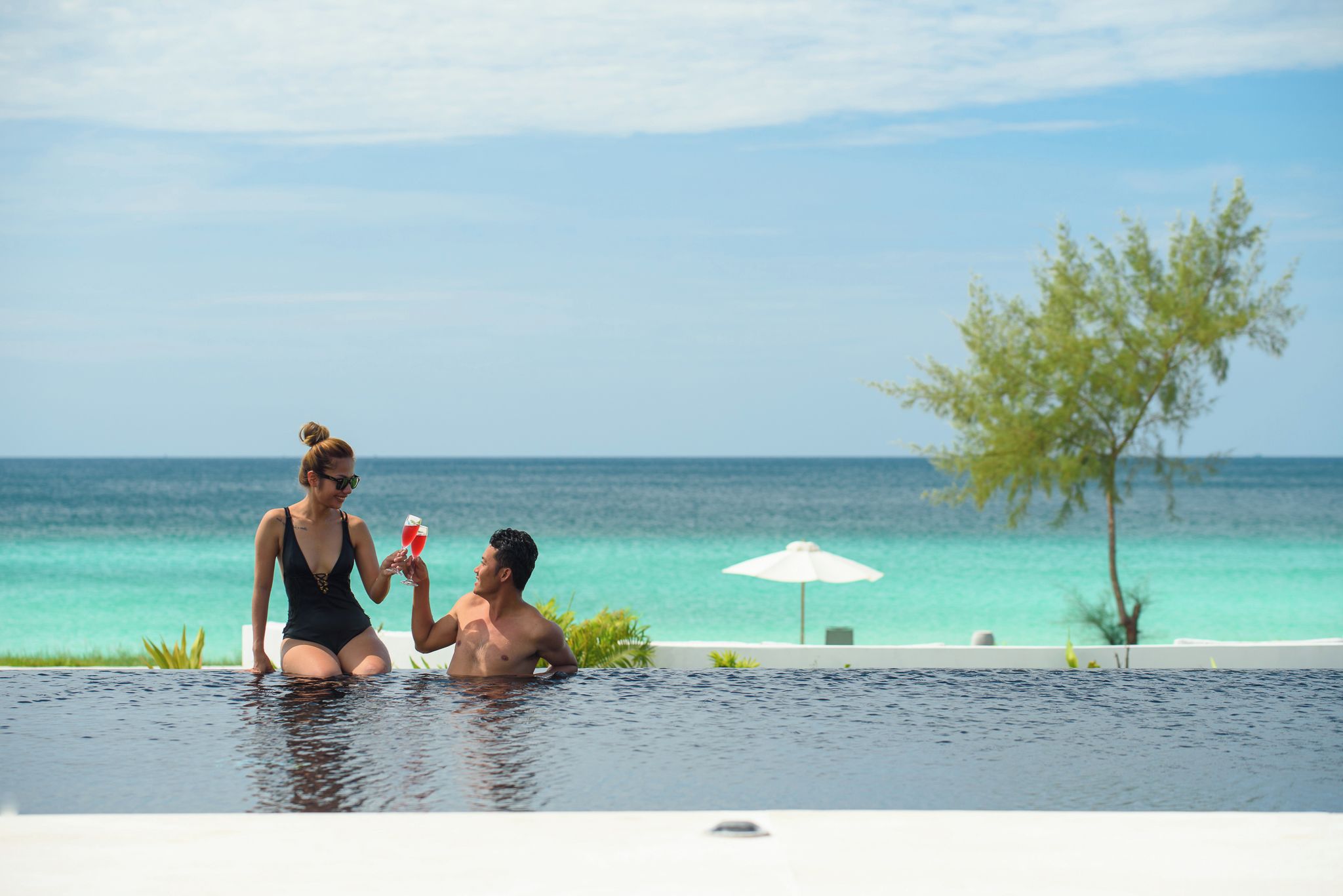 A couple in a swimming pool with a view of the ocean and a tree.