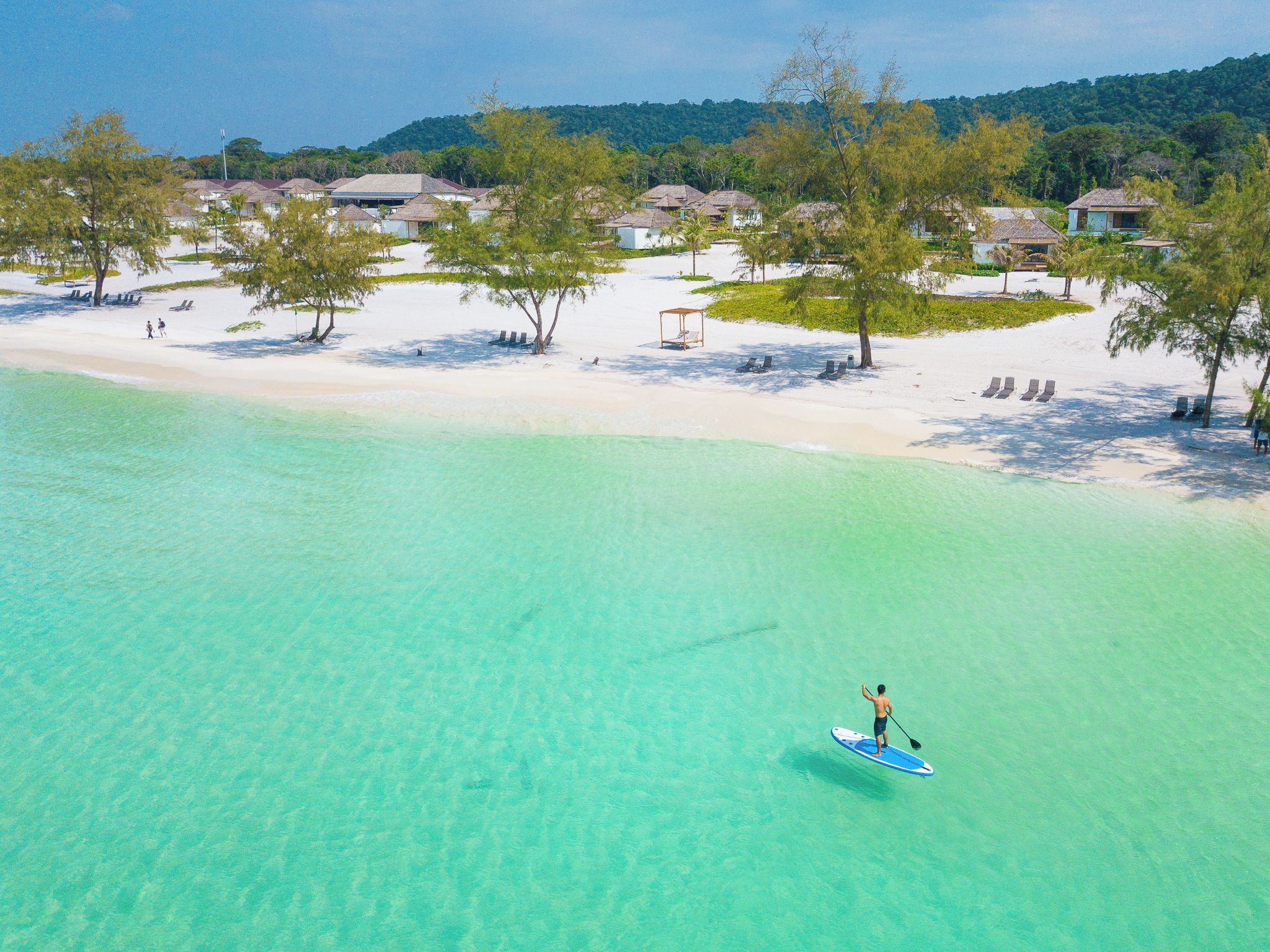 Una playa con agua turquesa cristalina, una persona haciendo paddleboard y una hilera de casas al fondo.