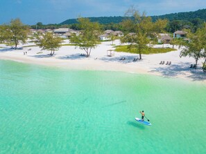 Una playa con agua turquesa cristalina, una persona haciendo paddleboard y una hilera de casas al fondo.