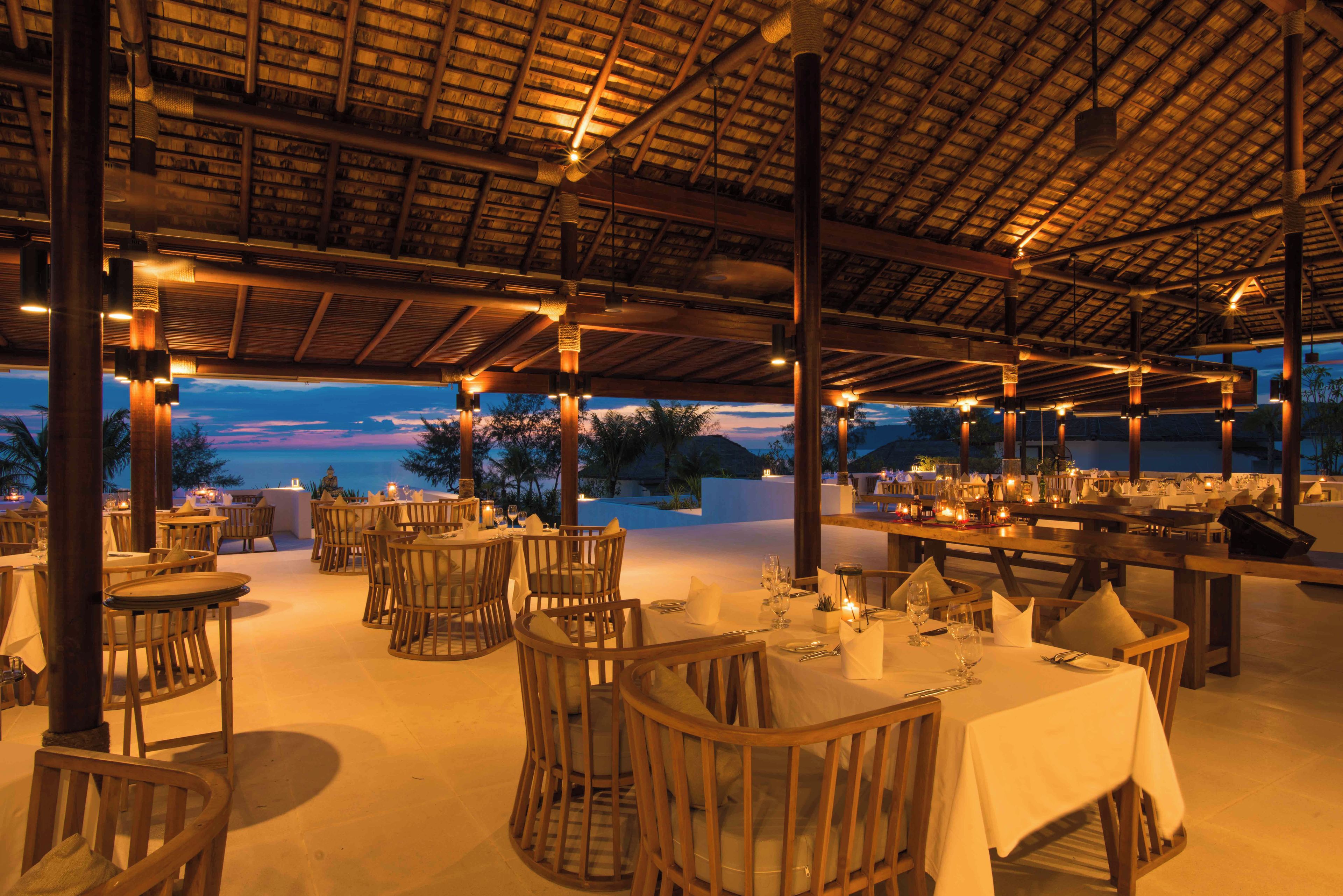 An outdoor dining area with tables and chairs, a thatched roof, and a view of the landscape.