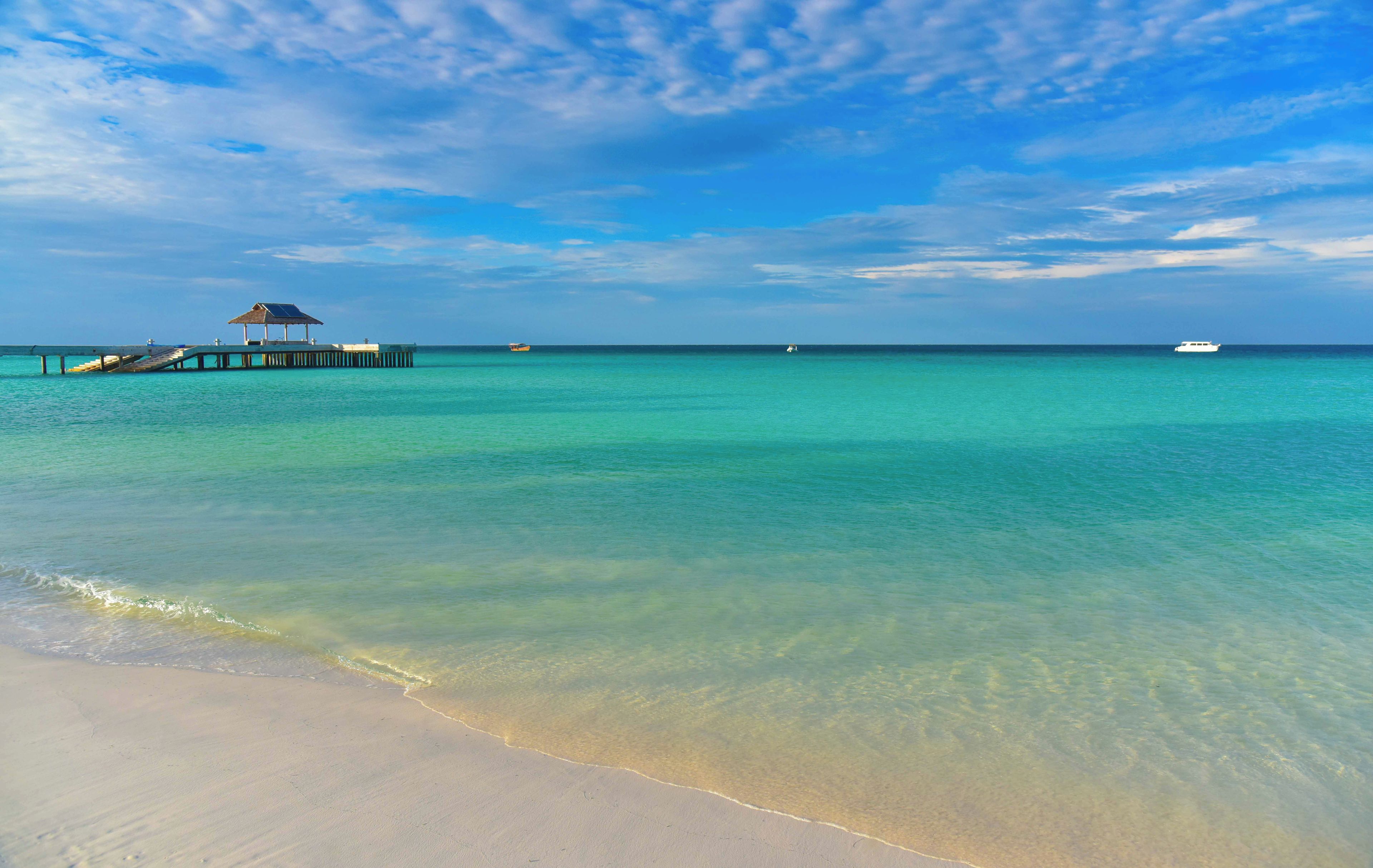 A beach with clear turquoise water, a pier with a small structure, and a boat in the distance.