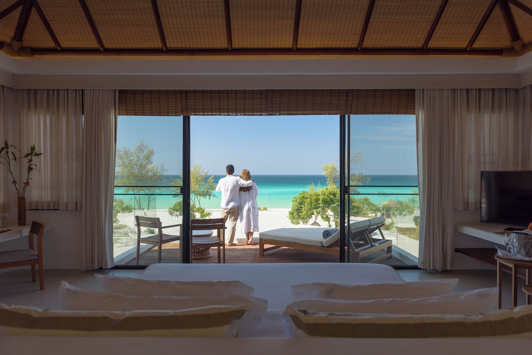 A hotel room with a large window, a bed, a TV, and a dining area with a view of the ocean.