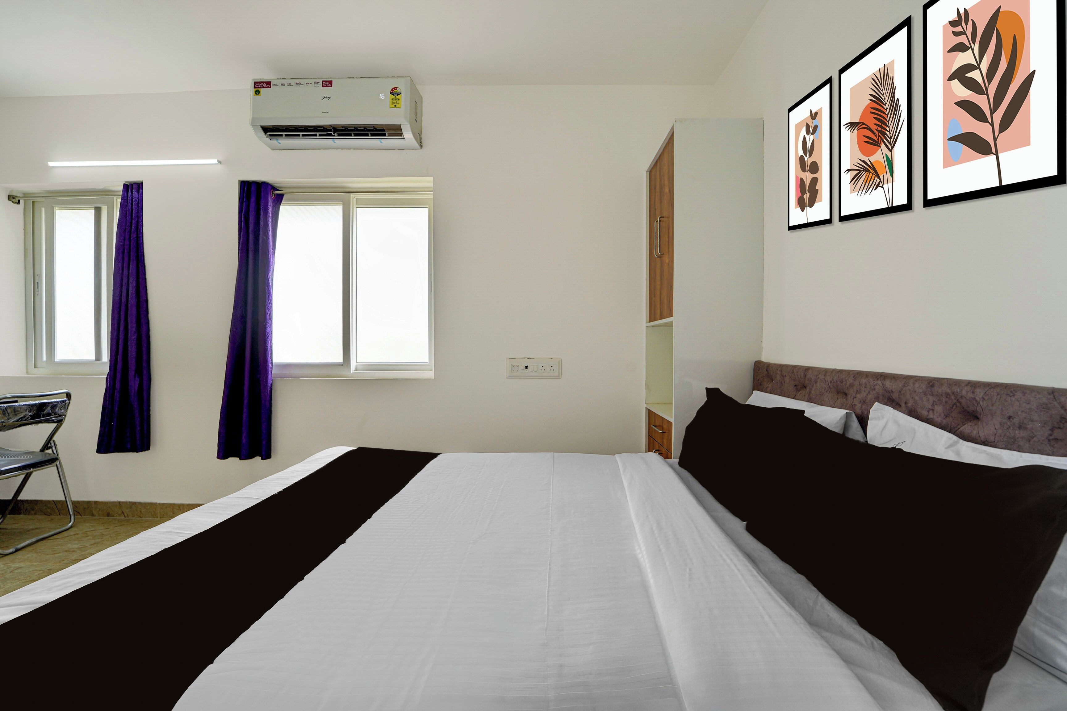 A hotel room with a bed, a desk, a chair, a window with curtains, an air conditioner, and framed artwork on the wall.
