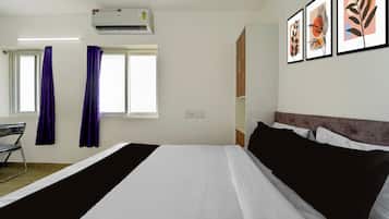 A hotel room with a bed, a desk, a chair, a window with curtains, an air conditioner, and framed artwork on the wall.
