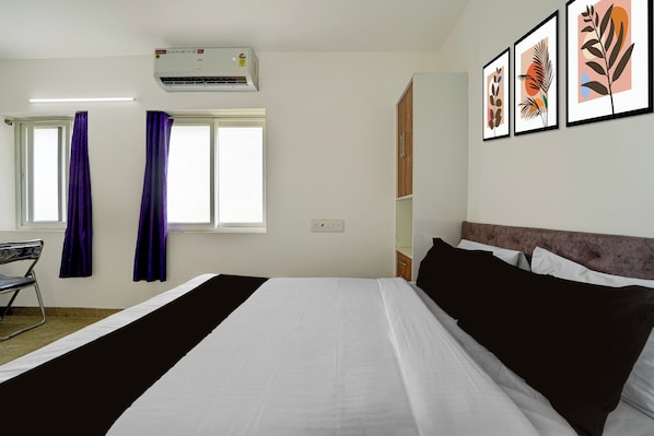 A hotel room with a bed, a desk, a chair, a window with curtains, an air conditioner, and framed artwork on the wall.
