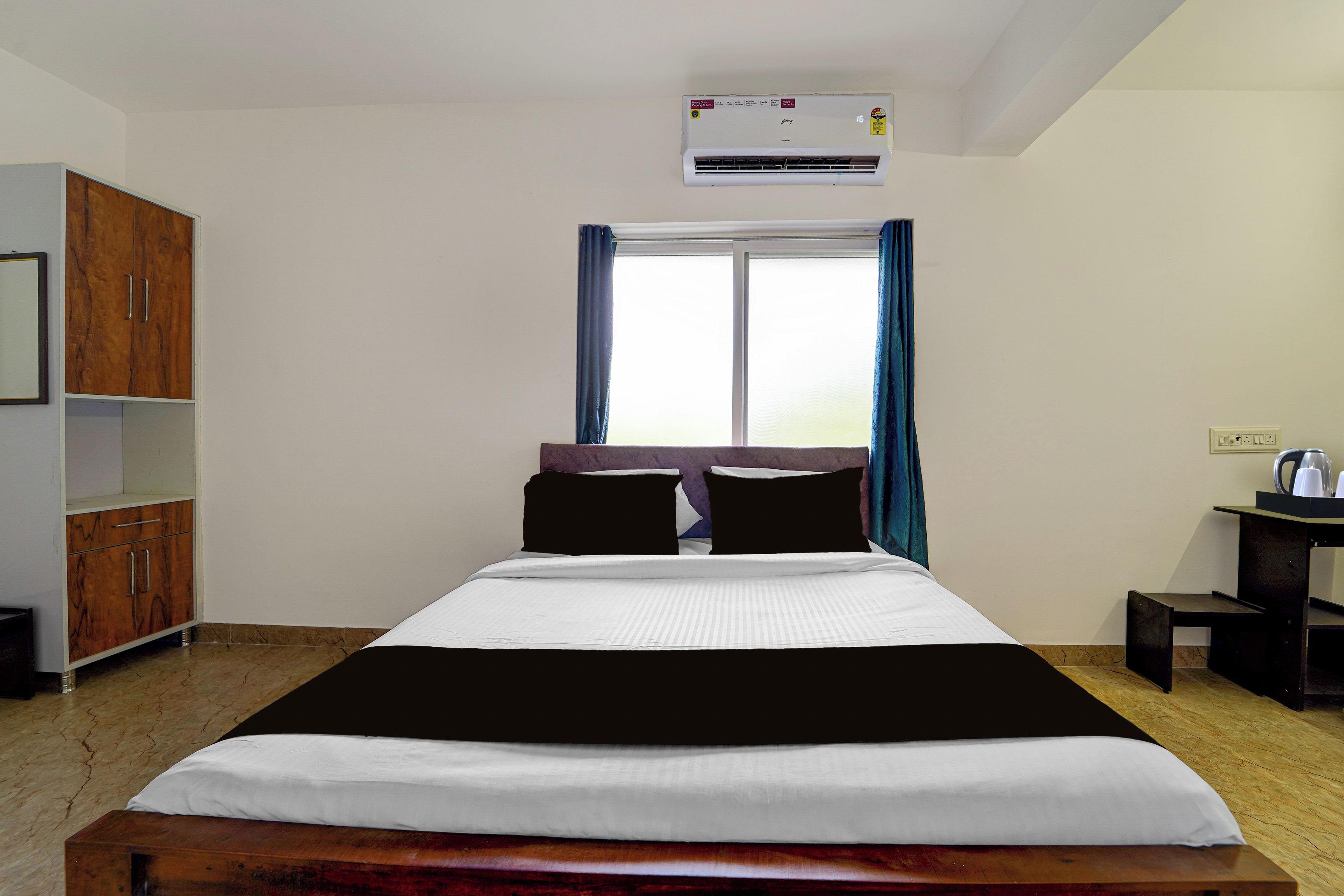 A hotel room with a bed, a wardrobe, a desk with a kettle, and an air conditioning unit.