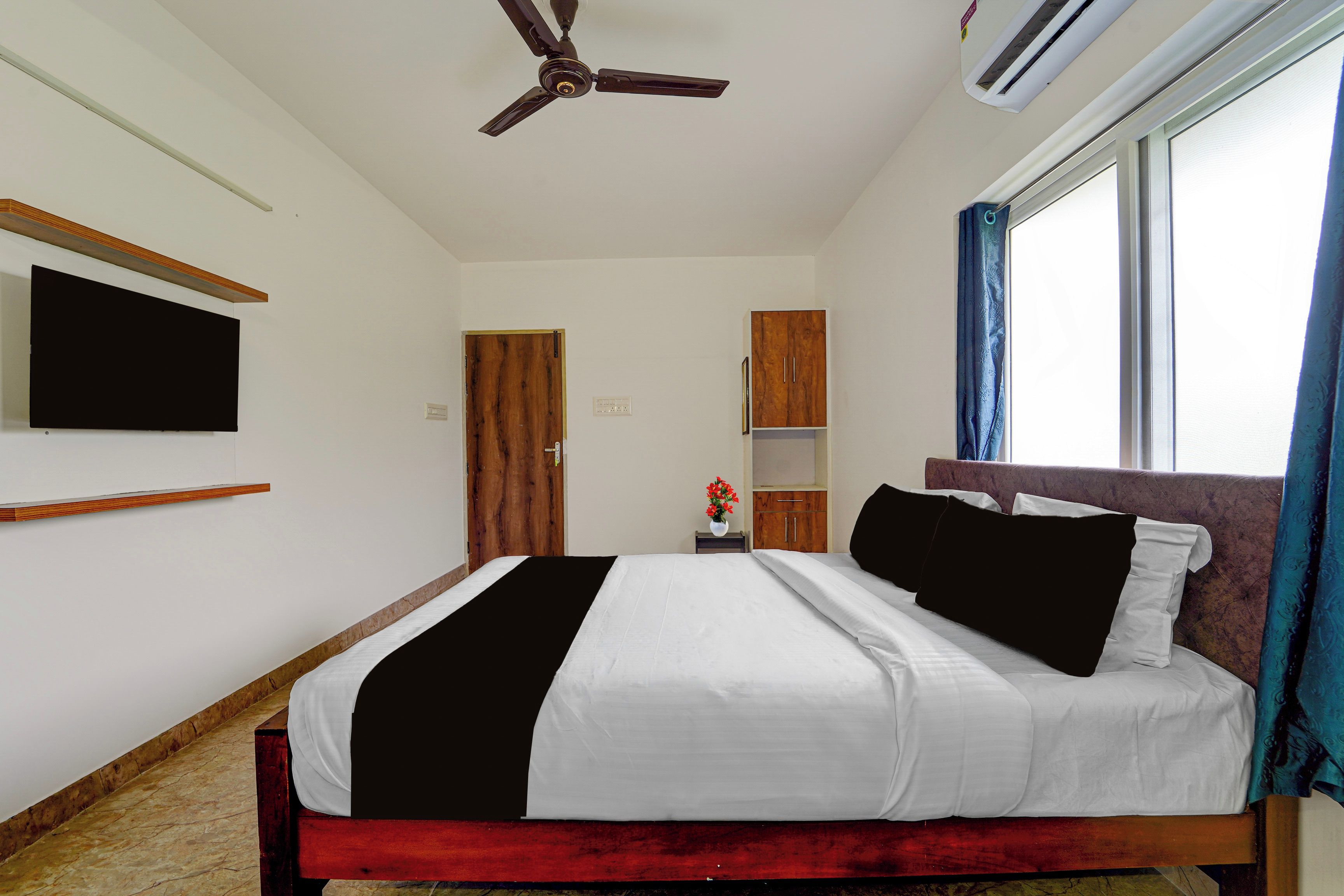 A bedroom with a bed, a ceiling fan, a TV mounted on the wall, a window with curtains, and a wooden door.