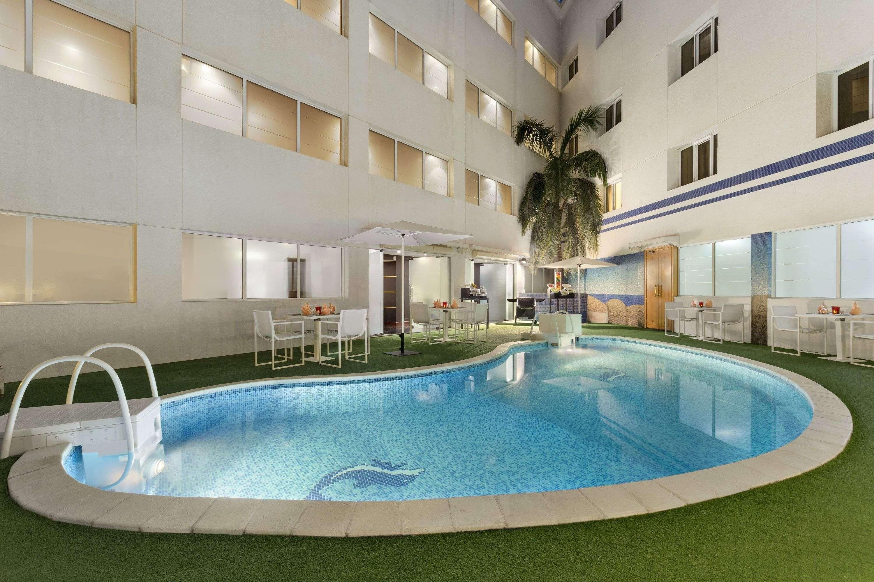An indoor swimming pool with artificial grass surrounding it, a seating area with tables and chairs, and a building with multiple windows and balconies.