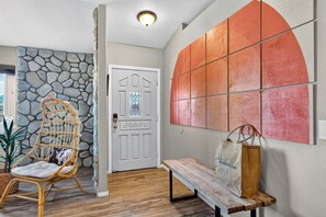 A hallway with a stone wall, a white door with a stained glass window, a wicker chair, a bench, and a wall with a large orange panel. - Rustic Ranch | Hot Tub + Fenced Yard | Stargazing + Desert Views | Rawhide Ranch (Pioneertown)