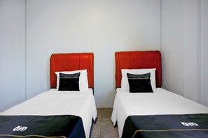 Dos camas individuales con cabeceros rojos y almohadas negras, sábanas blancas y una pared blanca lisa.
