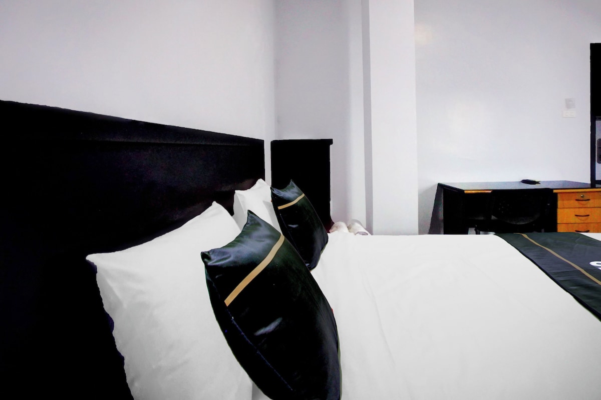 Una cama con ropa de cama blanco y negro, un cabecero negro y una mesita de noche pequeña con una lámpara.