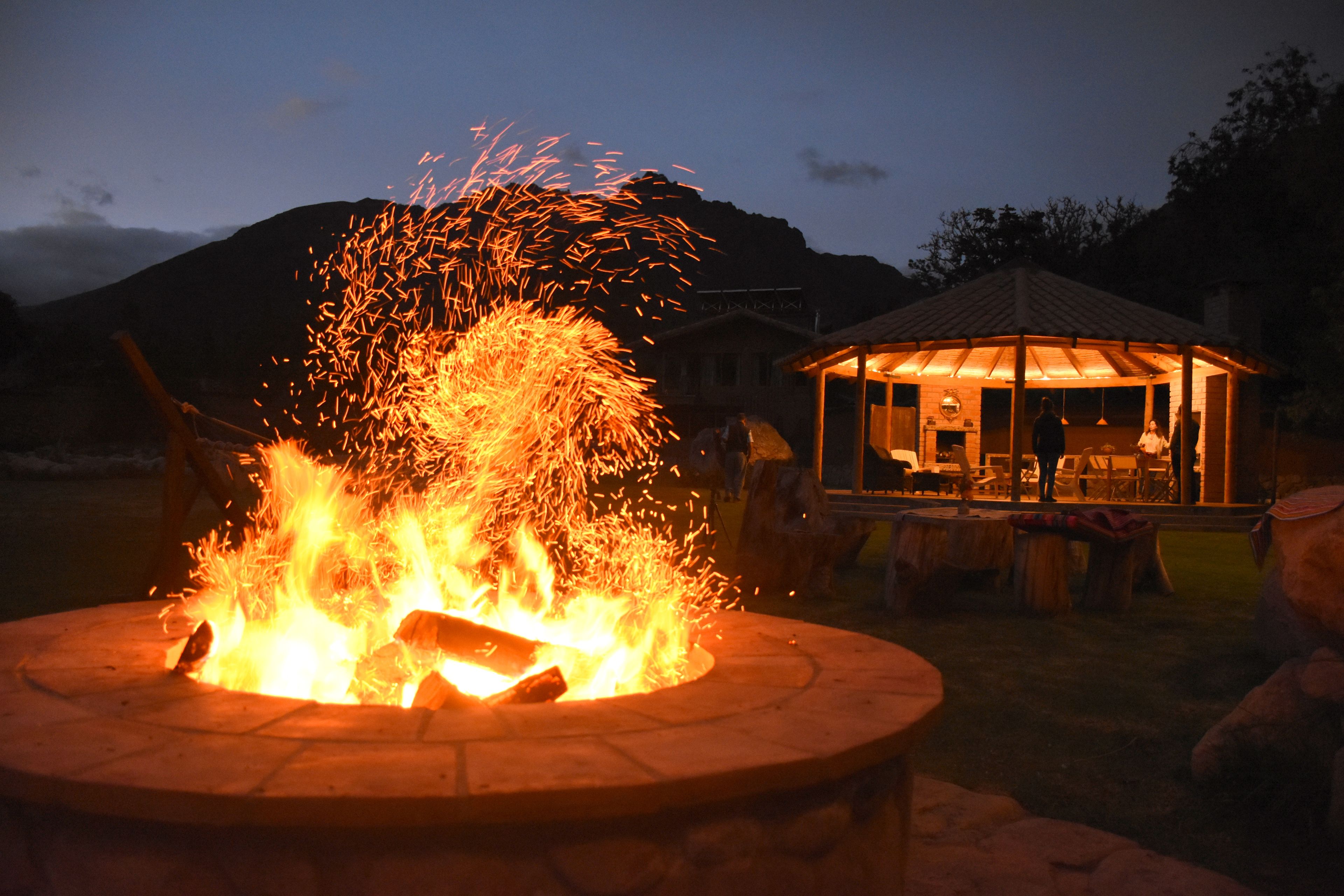A circular fire pit with visible flames and sparks, surrounded by a covered seating area with tables and chairs.