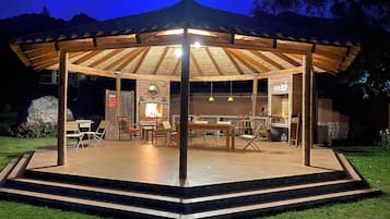 A covered outdoor seating area with a wooden roof, tables, chairs, and a brick wall.