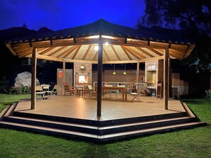 Un espacio de estar al aire libre techado, con techo de madera, mesas, sillas y pared de ladrillo.