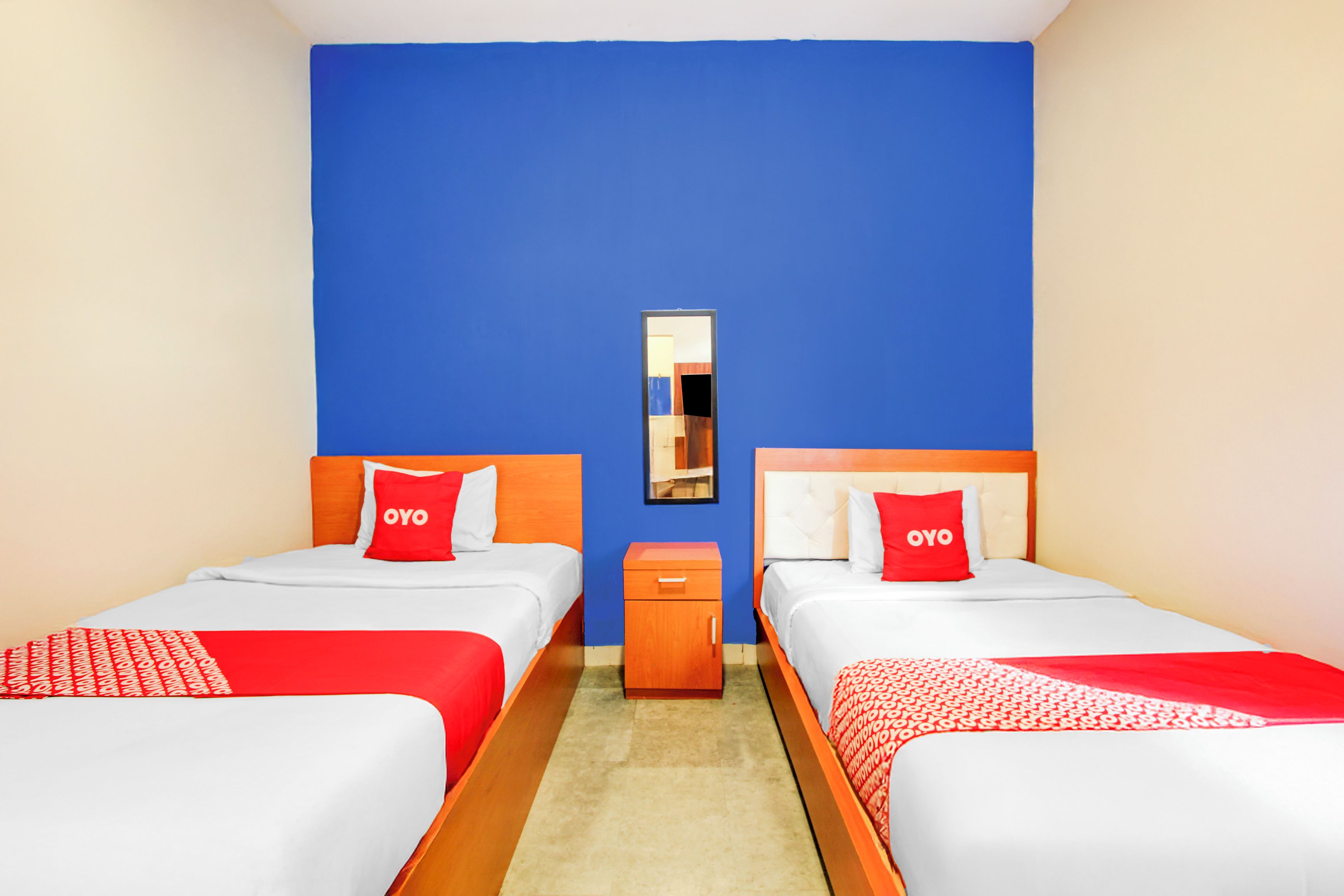 Habitación de hotel con dos camas, cada una con colcha y almohadas rojas y blancas, una mesita de noche de madera y un espejo en la pared.