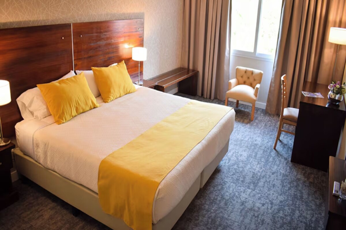 Habitación de hotel con una cama grande, dos mesitas de noche, una silla, un escritorio y un ventanal con cortinas.