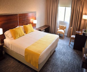 Habitación de hotel con una cama grande, dos mesitas de noche, una silla, un escritorio y un ventanal con cortinas.