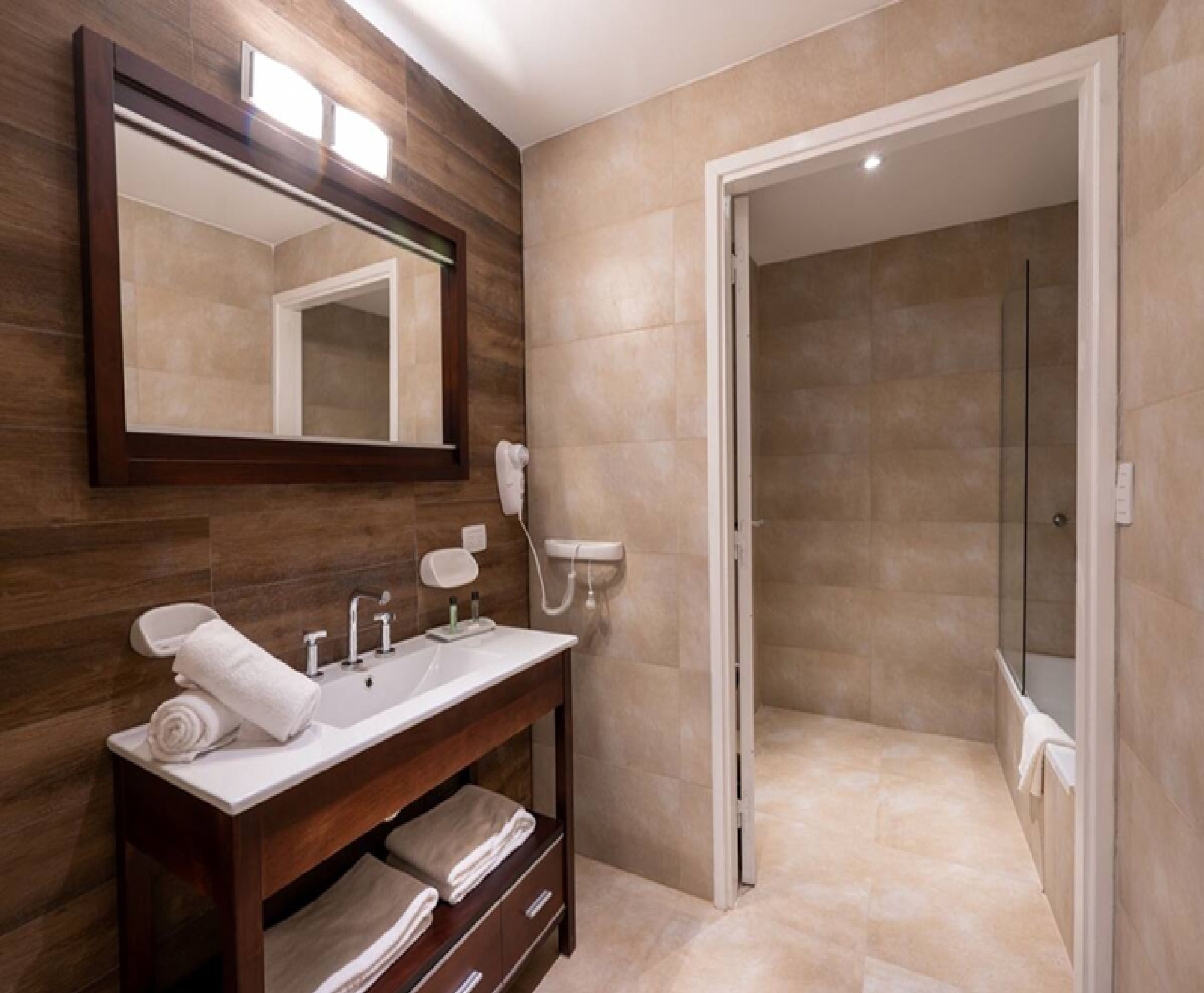 Un baño moderno con tocador de madera, un espejo grande, ducha y azulejos color beige.