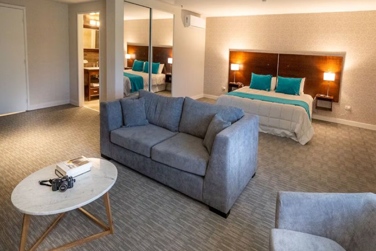 Habitación de hotel con sofá gris, una mesa redonda, cama con ropa de cama color turquesa y mesita de noche con lámpara.