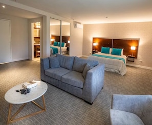 Habitación de hotel con sofá gris, una mesa redonda, cama con ropa de cama color turquesa y mesita de noche con lámpara.