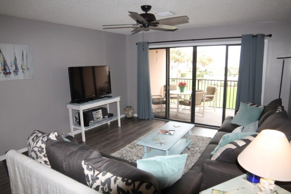 A living room with a sofa, coffee table, TV, and a balcony with outdoor furniture.